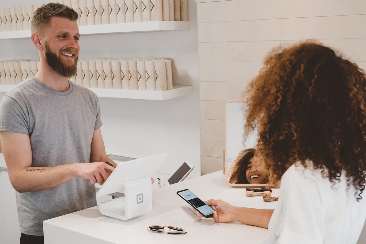 Un commerçant souriant échange avec une cliente au comptoir de sa boutique, illustrant la relation de confiance en magasin.